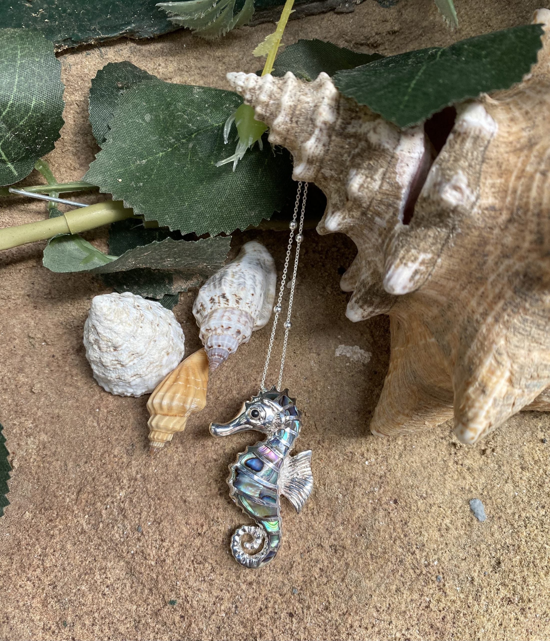 Silver seahorse necklace on sand with shells and leaves