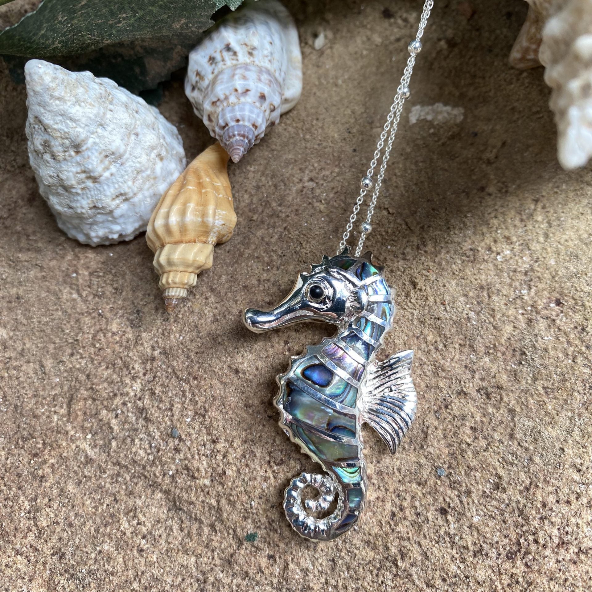 Silver seahorse necklace on sand with shells and leaves
