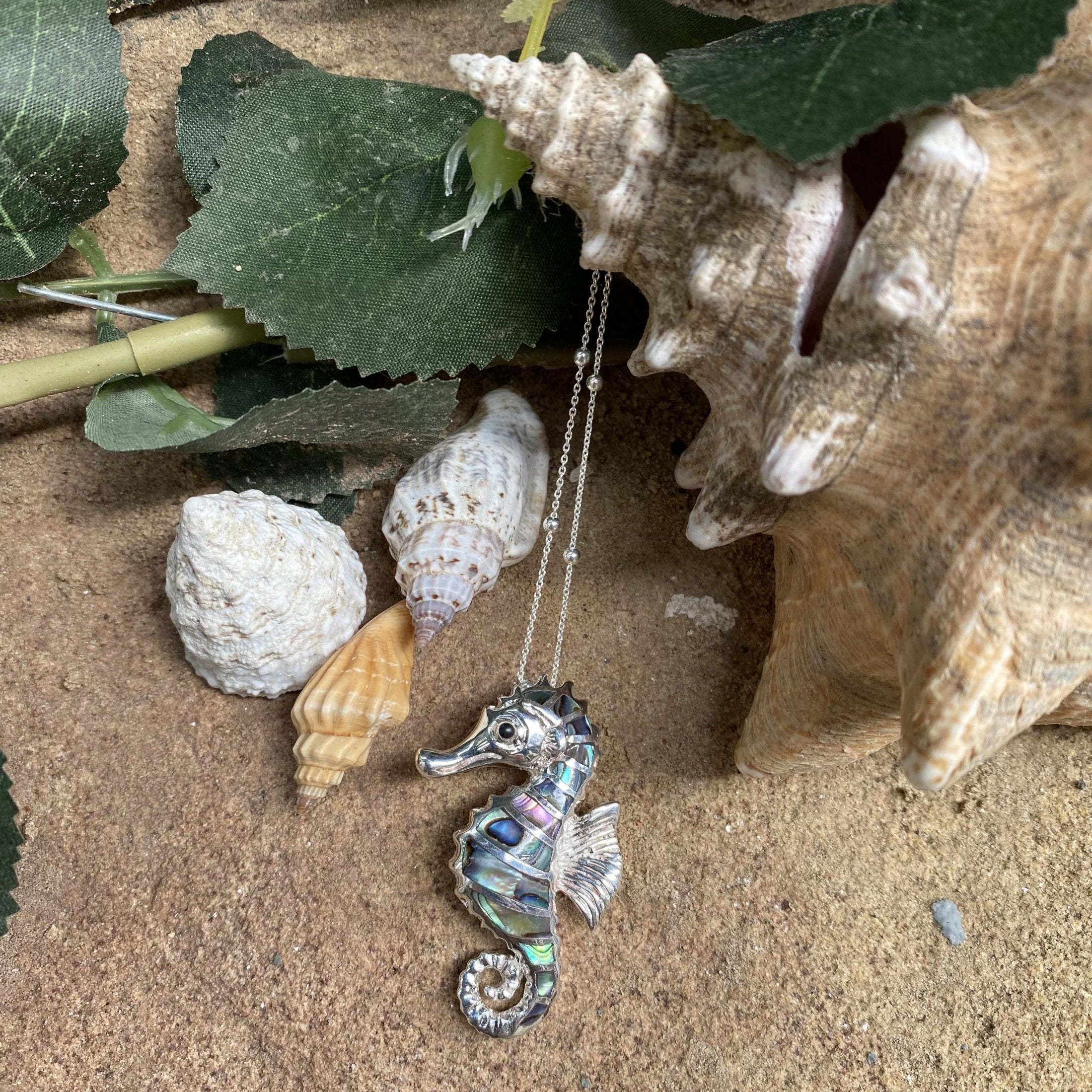 Silver seahorse necklace on sand with shells and leaves