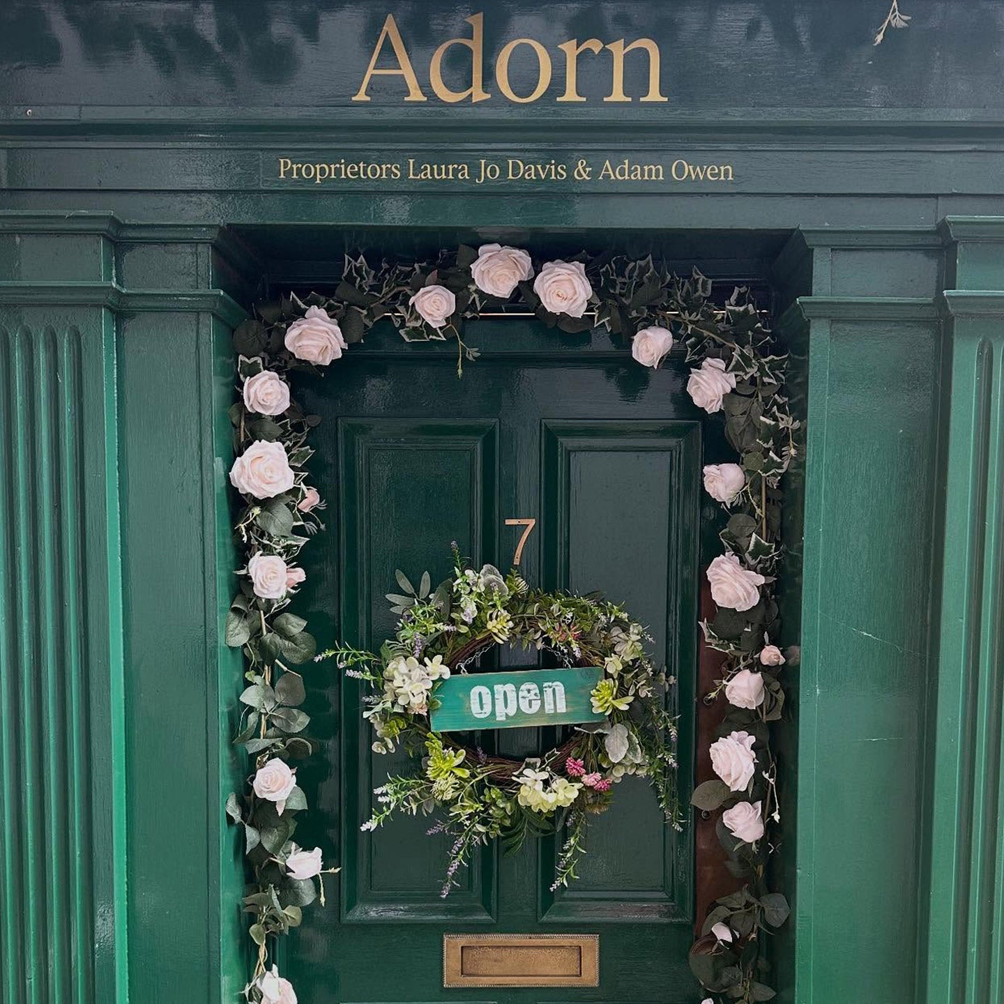Decorative wreath with flowers and a sign on a green door, featuring the brand 'Adorn'.