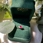 A gold ring with an oval red gemstone displayed in an open dark green velvet ring box, surrounded by white flowers and green leaves.