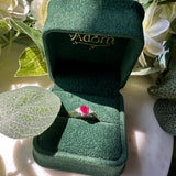 A gold ring with an oval red gemstone displayed in an open dark green velvet ring box, surrounded by white flowers and green leaves.