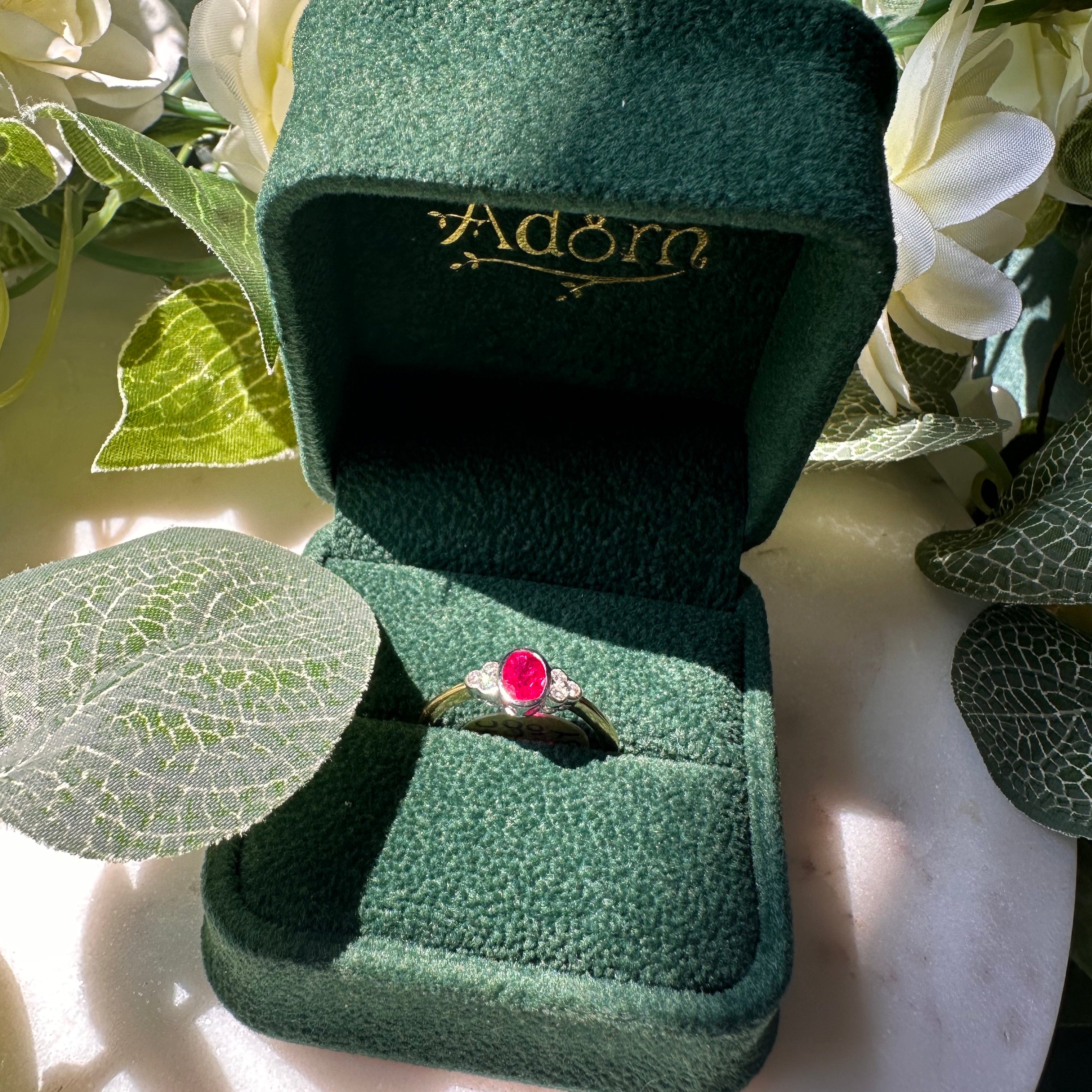 A gold ring with an oval red gemstone displayed in an open dark green velvet ring box, surrounded by white flowers and green leaves.