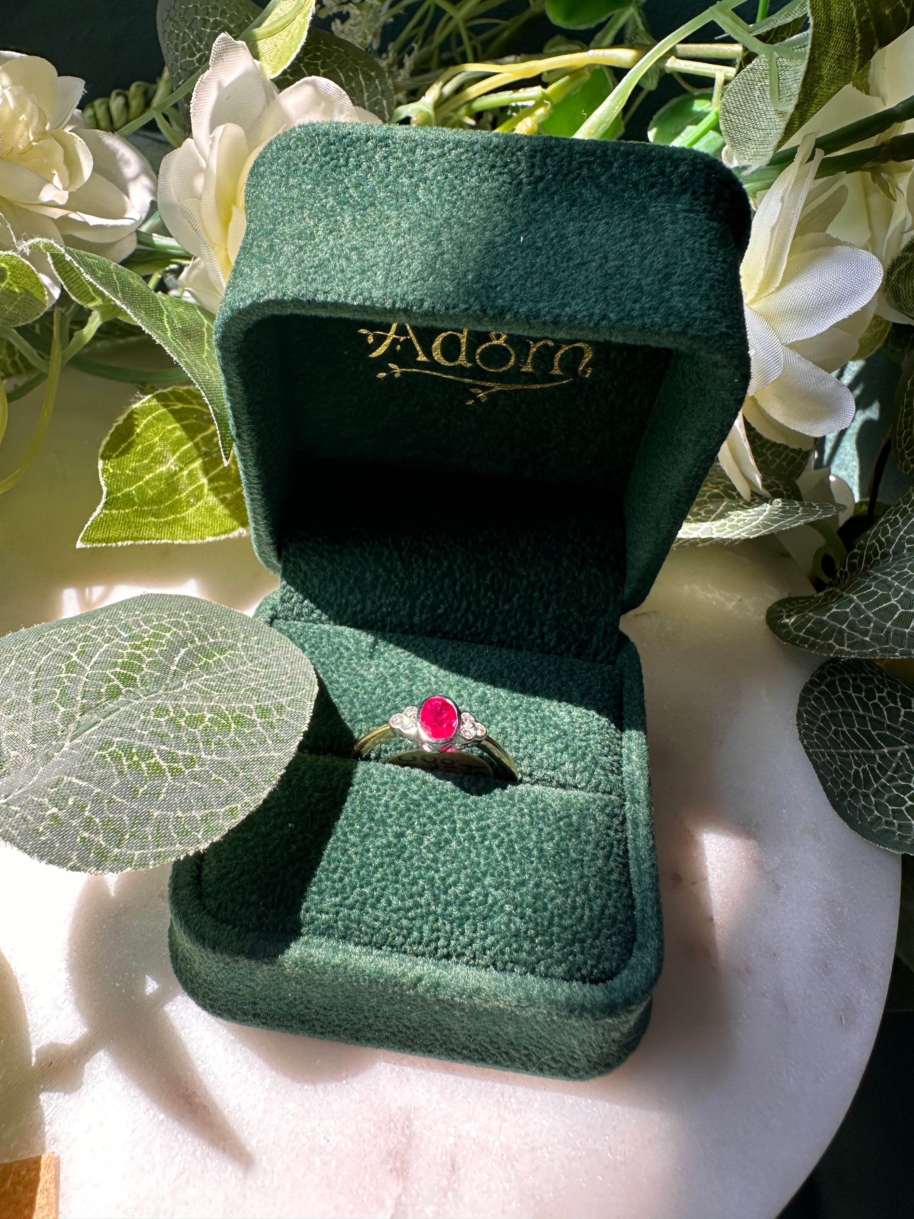 A gold ring with an oval red gemstone displayed in an open dark green velvet ring box, surrounded by white flowers and green leaves.