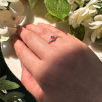 Laura Jo's hand wearing a ring with a round red centre gemstone and small diamonds photographed among white flowers and green leaves.