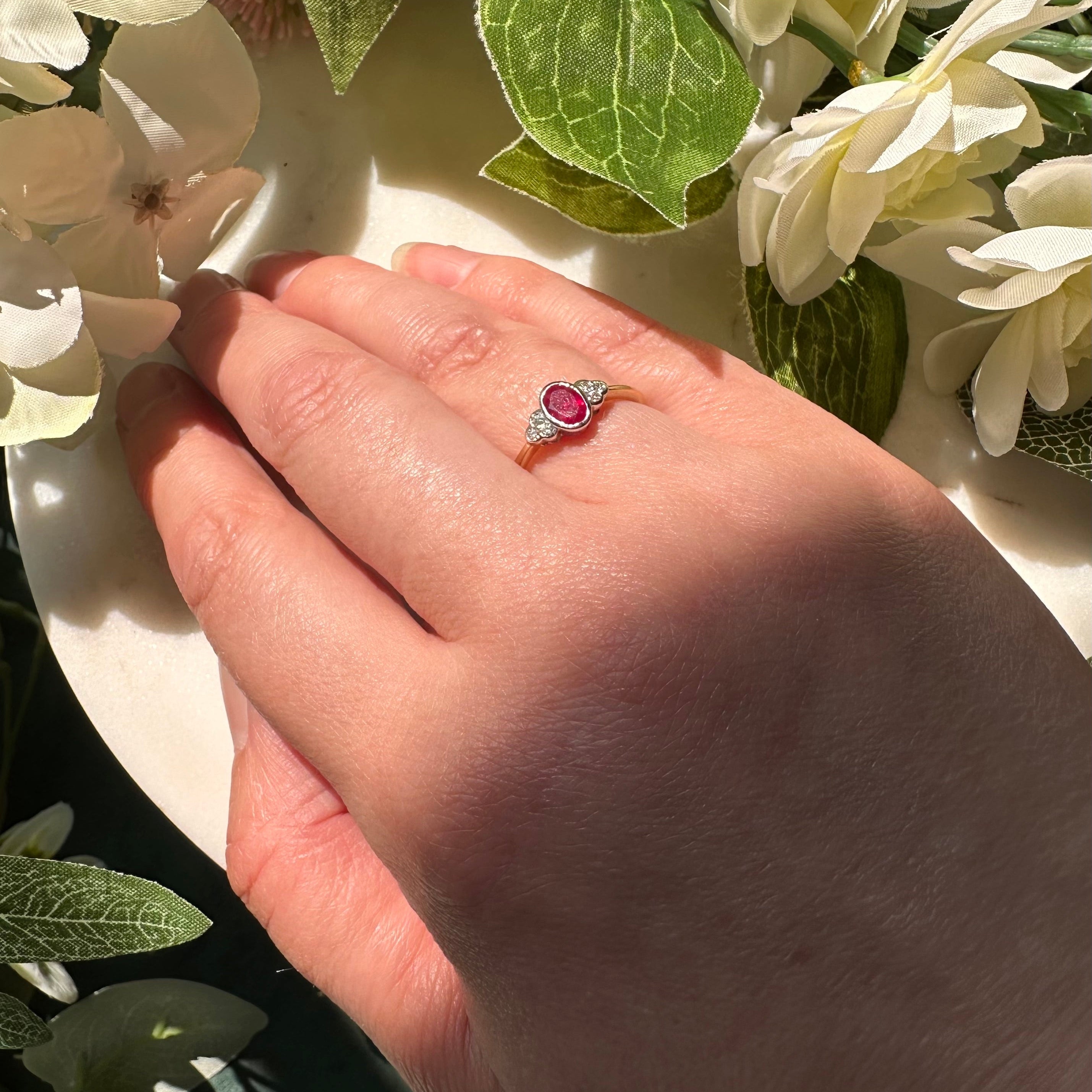 Laura Jo's hand wearing a ring with a round red centre gemstone and small diamonds photographed among white flowers and green leaves.
