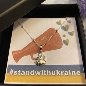 A silver peace dove pendant in a black box with a drawing of a megaphone with blue and yellow hearts coming out . Underneath is a hashtag which says #standwithukraine