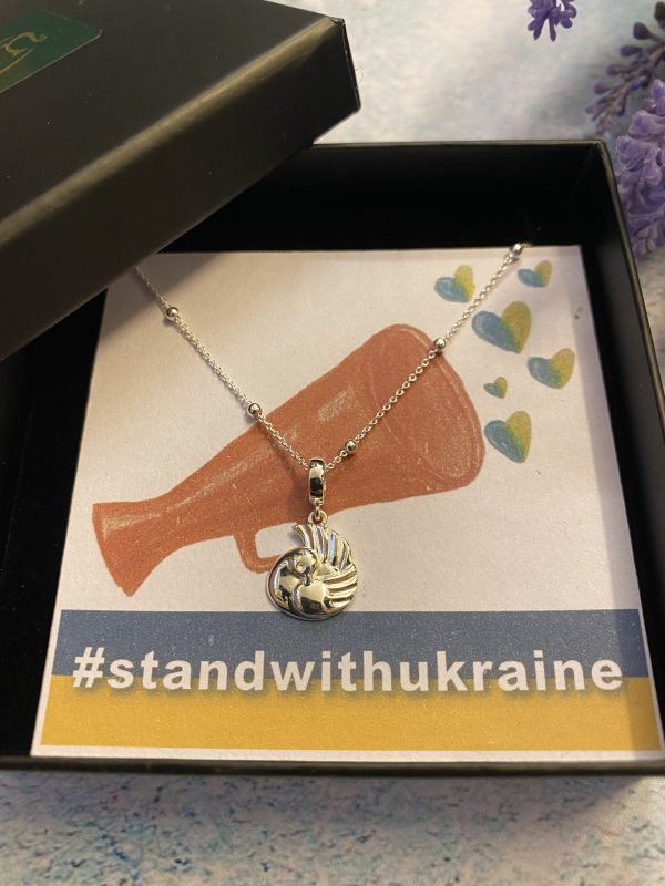 A silver peace dove pendant in a black box with a drawing of a megaphone with blue and yellow hearts coming out . Underneath is a hashtag which says #standwithukraine